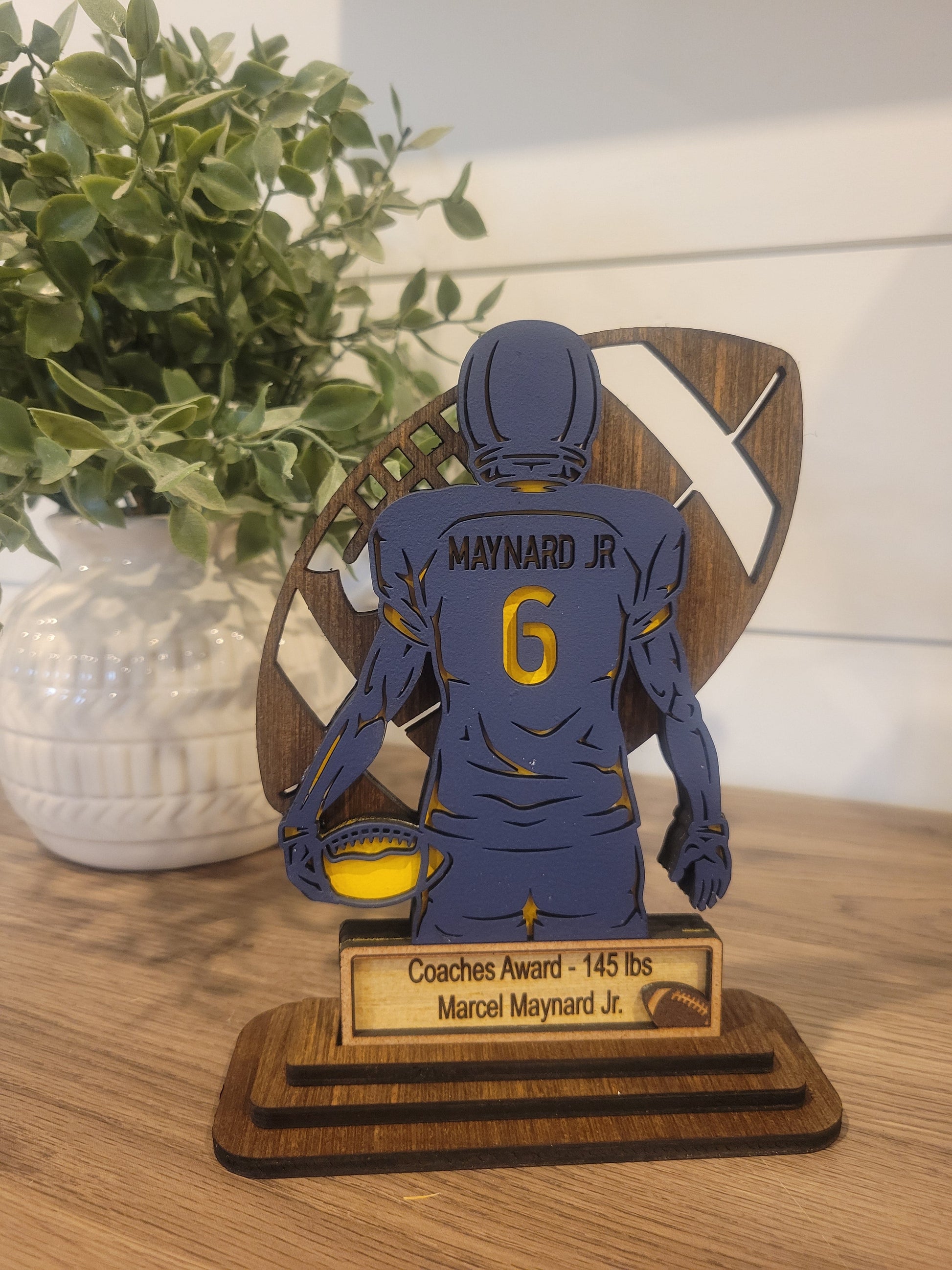 A custom youth football team trophy award featuring a wood sign with a player in a blue jersey holding a football, with the number 6 on it. The sign also includes a plaque with the text 'COACH AWARD MARJORIE JR 145 LBS'.
