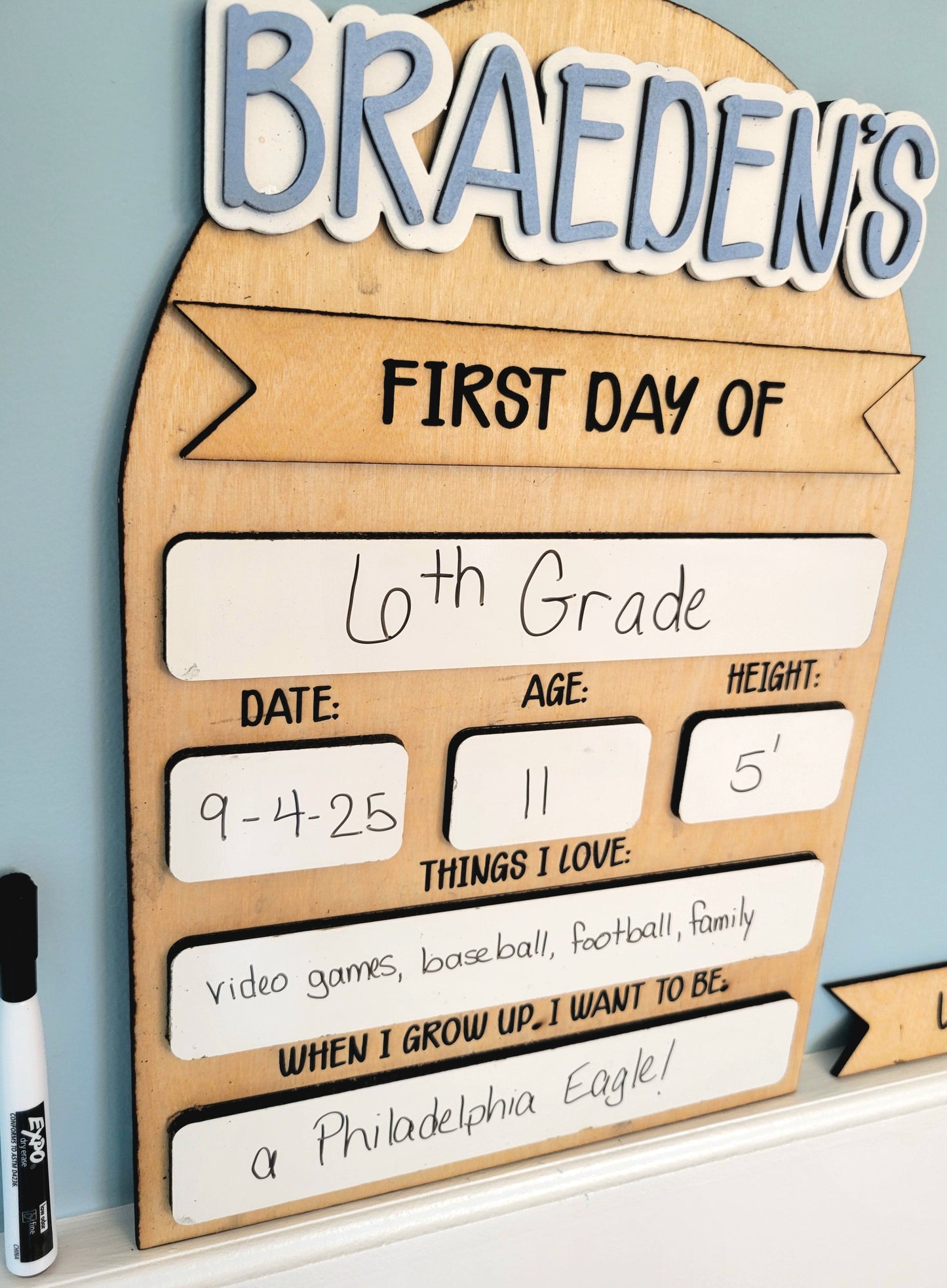 Wooden sign with 'Braeden's First Day of 6th Grade' details on a light blue wall.