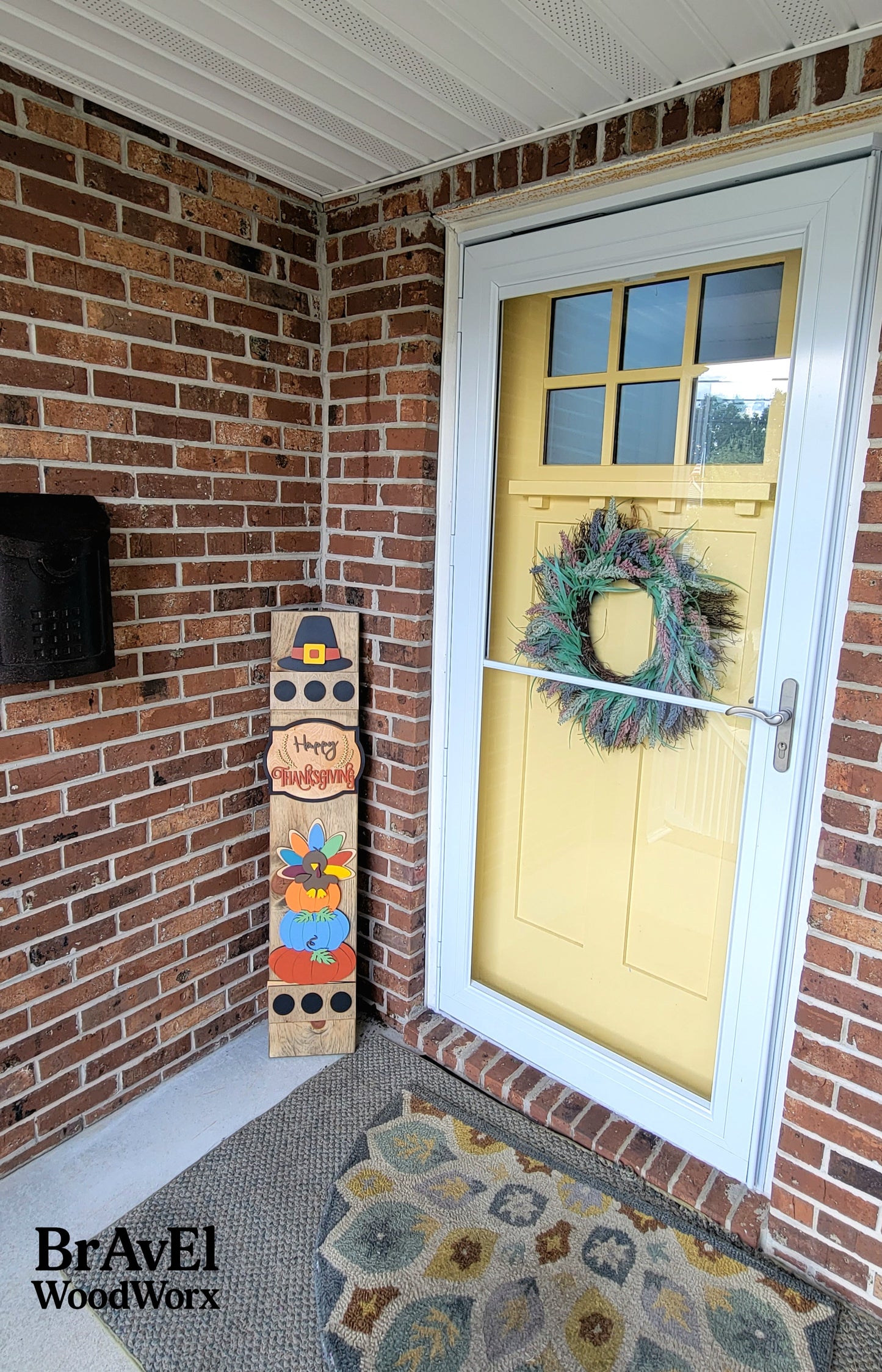 Front door with a wreath, decorative sign, and BravEl WoodWorx branding on a brick wall.