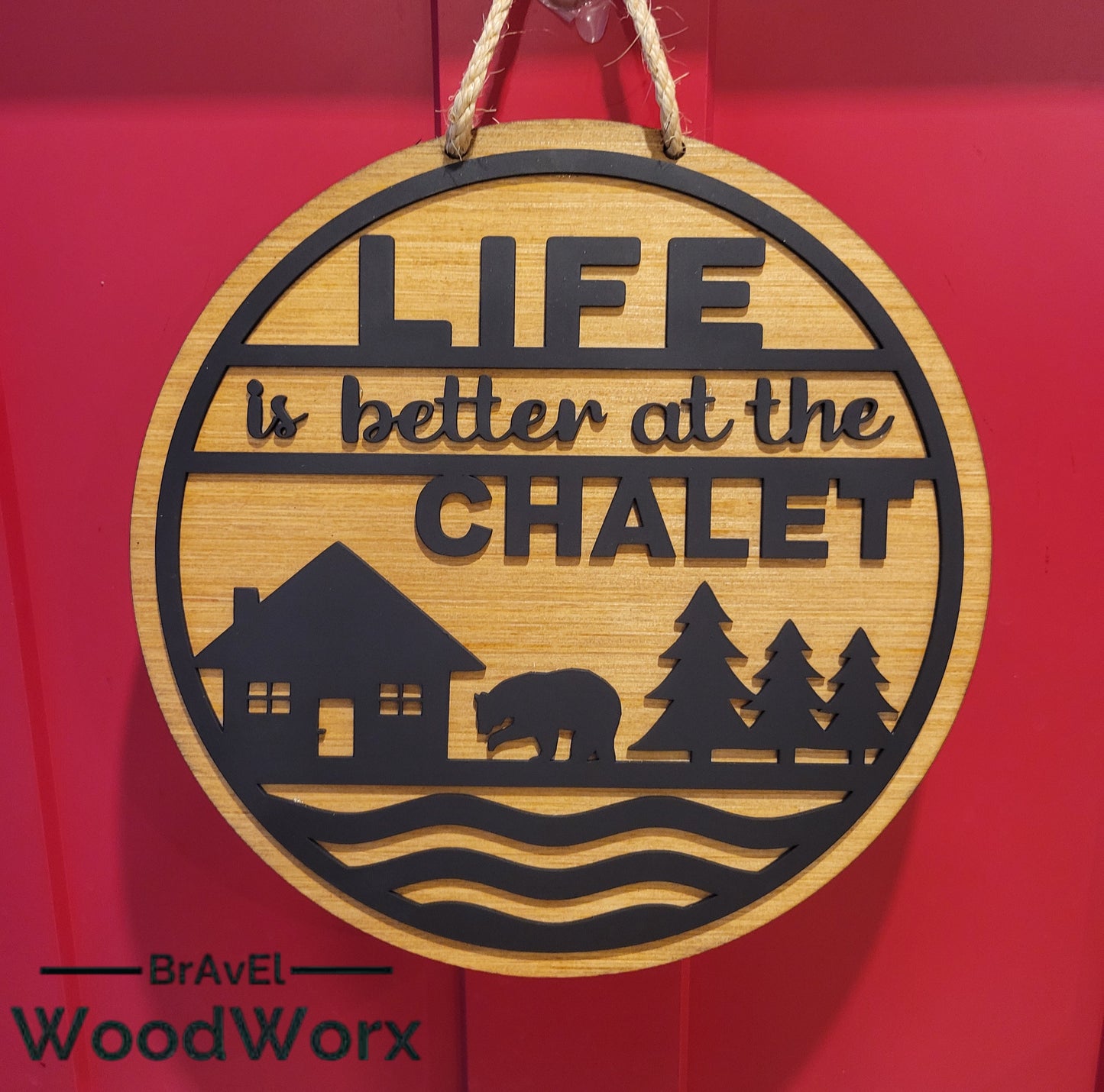 A round wooden sign with the text 'Life is better at the Chalet' carved on it, alongside an image of a house with a bear and trees. The sign has a darker finish around the text and a lighter finish on the background.
