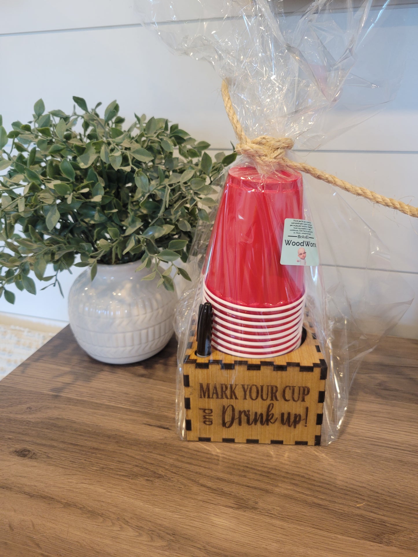 A set of red cup holders with a personalized sharpie cup holder, packaged in a clear plastic bag, tied at the top with a twine. The cup holders are displayed on a wooden surface with a white vase and green foliage in the background.