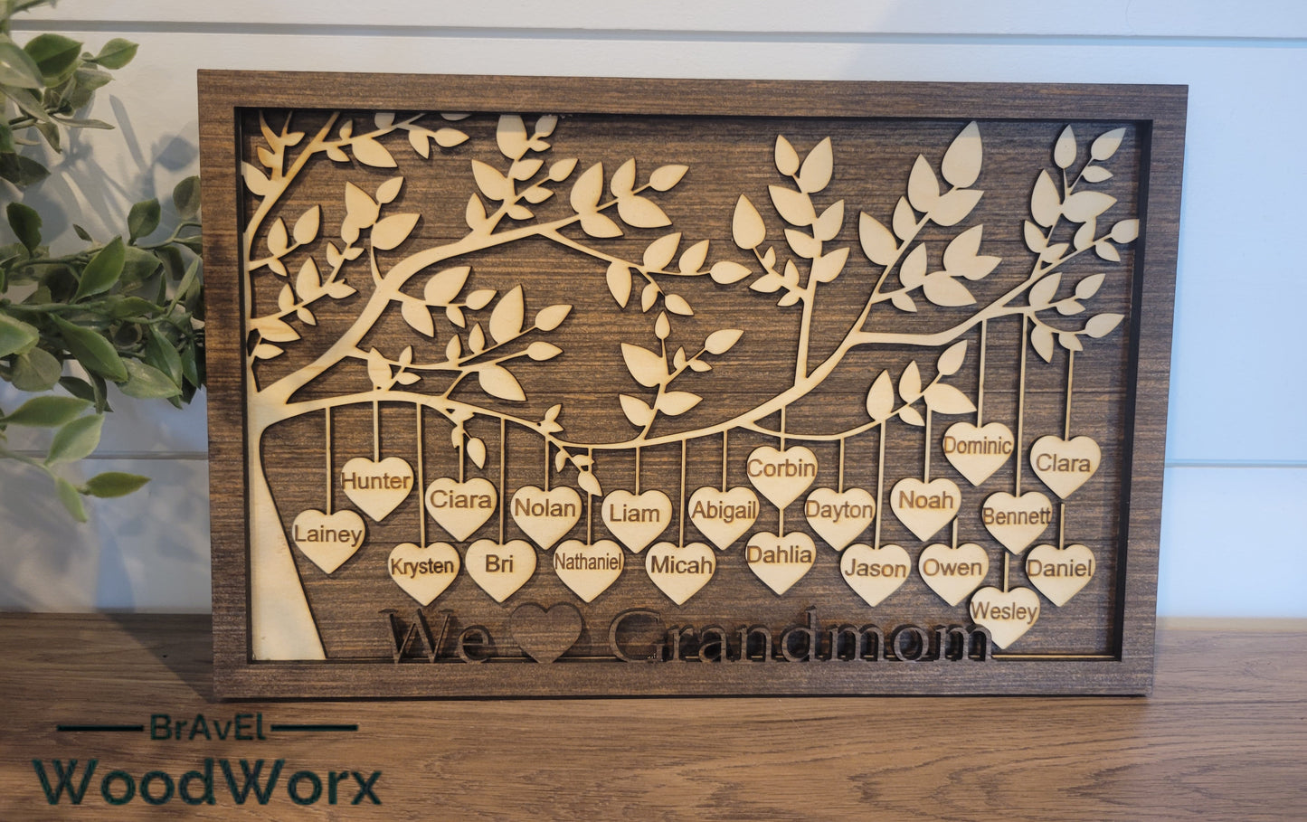 A personalized wooden family tree sign with a layered laser-cut tree design and heart name tags, stained wood background, and the text 'We Love Grandmom'.