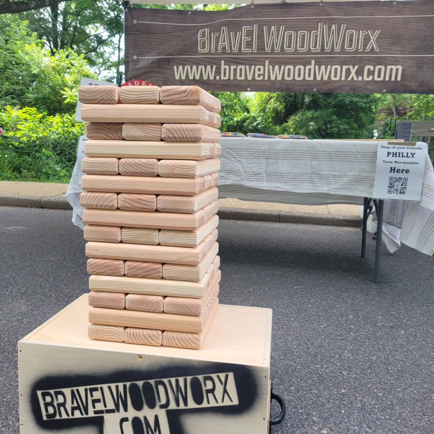 A wooden stack of blocks on a cardboard box with 'BrAvEl WoodWorx' printed on it, set outdoors with a 'Giant Block Rental Day' sign in the background.