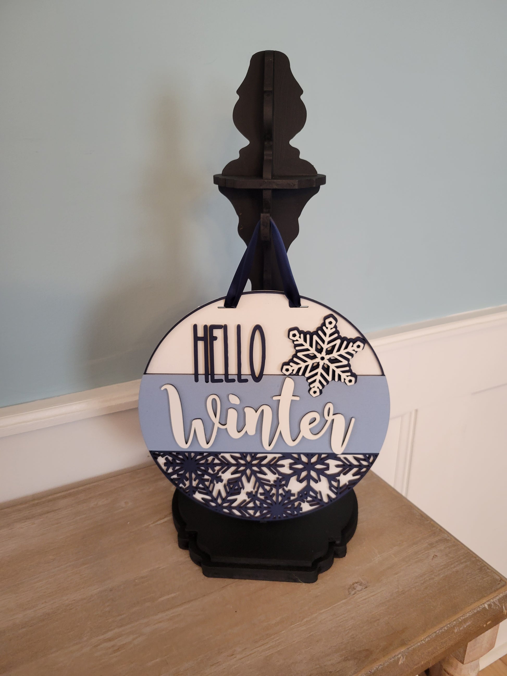 A wooden "Hello Winter" snowflake shelf stand featuring an intricate laser-cut snowflake design and the words "Hello Winter" in stylish script. Painted in a festive winter-themed color palette, it sits beautifully on a mantel or shelf, adding a cozy seasonal touch to your home decor. classic stand
