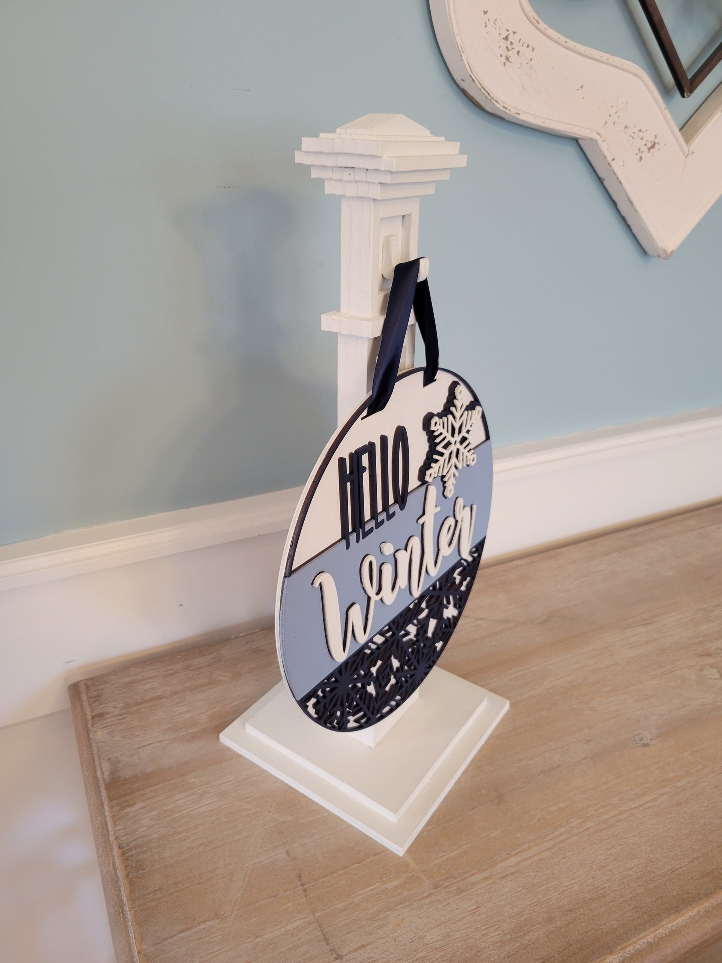 A wooden "Hello Winter" snowflake shelf stand featuring an intricate laser-cut snowflake design and the words "Hello Winter" in stylish script. Painted in a festive winter-themed color palette, it sits beautifully on a mantel or shelf, adding a cozy seasonal touch to your home decor. side view
