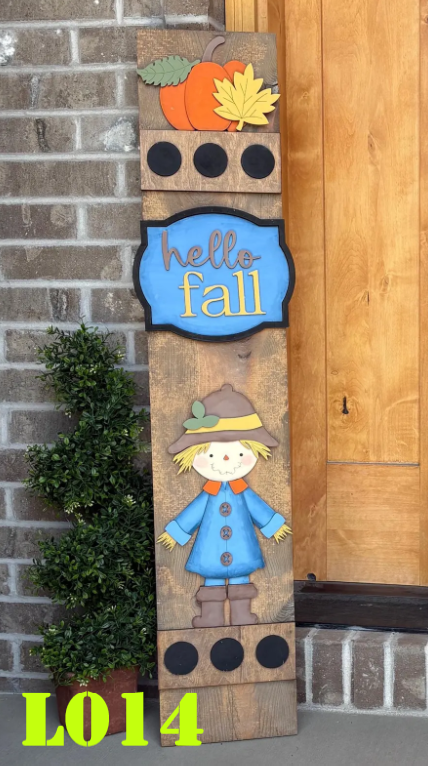 Decorative fall-themed wooden sign with a scarecrow, pumpkins, and leaves against a brick wall.