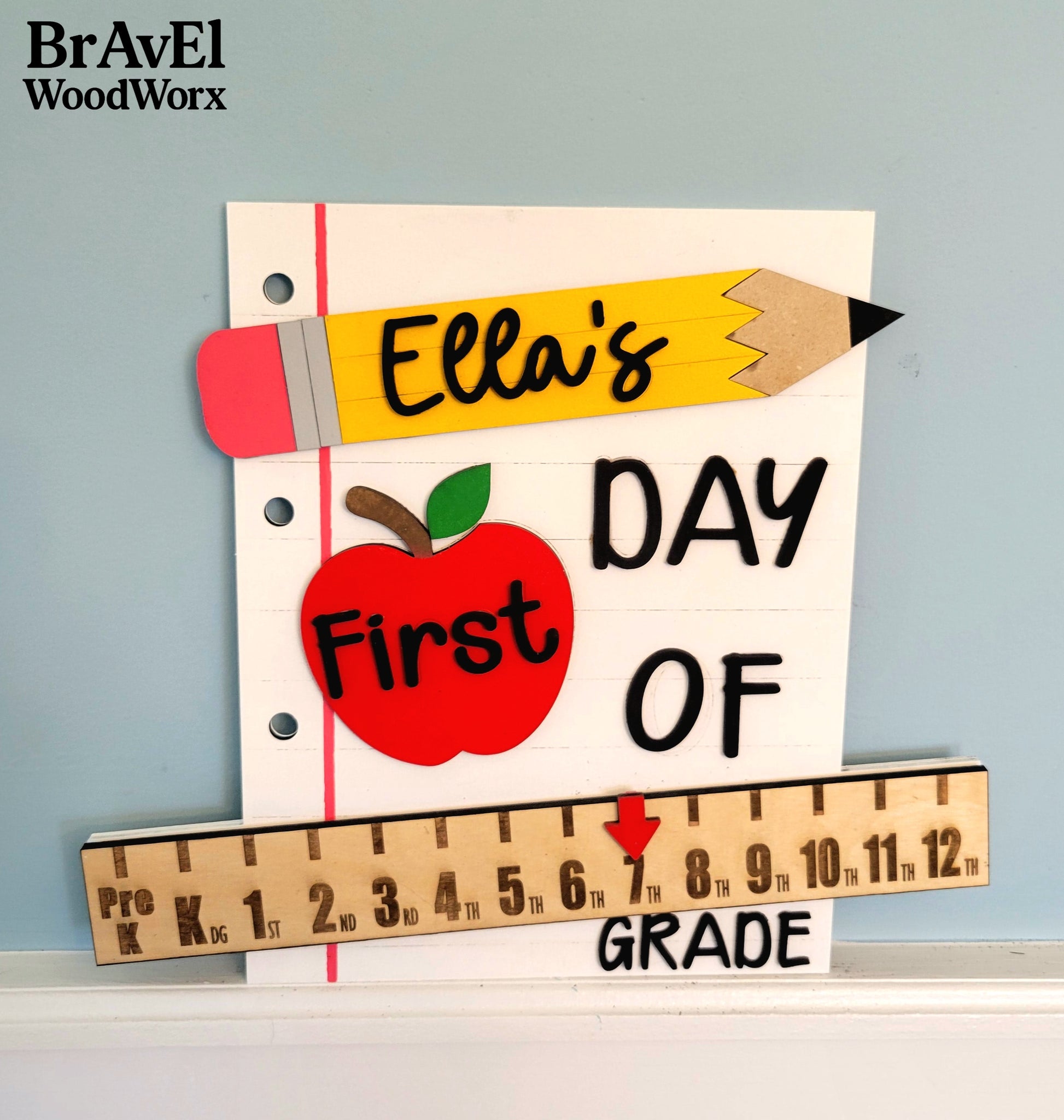 Decorative sign for 'Ella's First Day of Grade' with a pencil, apple, and ruler design on a light blue background.