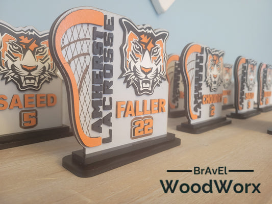 A series of personalized wooden lacrosse player shelf sitters with bases, each featuring a different name and number, arranged on a table.