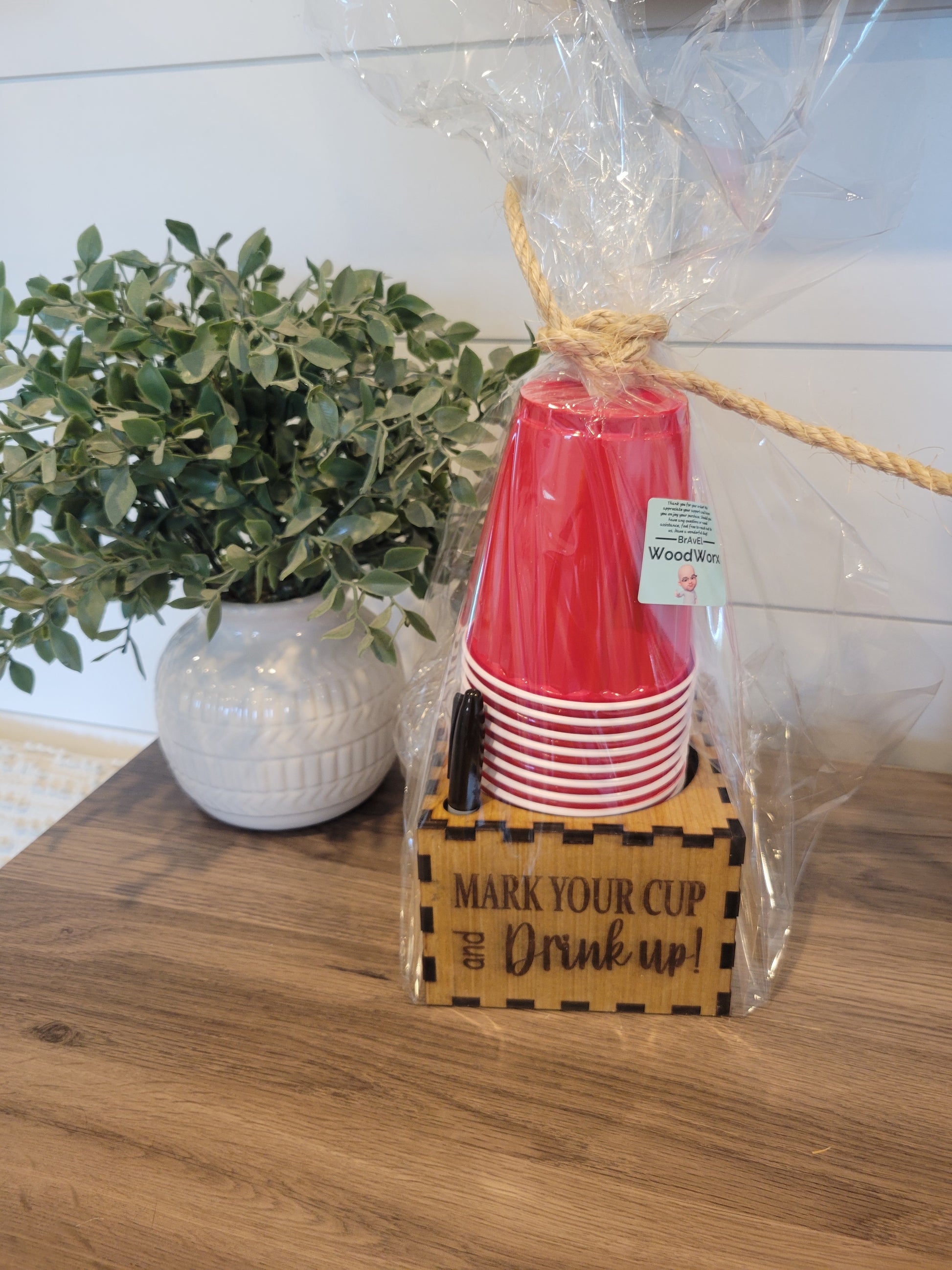 A set of red cup holders with a personalized sharpie cup holder, packaged in a clear plastic bag, tied at the top with a twine. The cup holders are displayed on a wooden surface with a white vase and green foliage in the background.