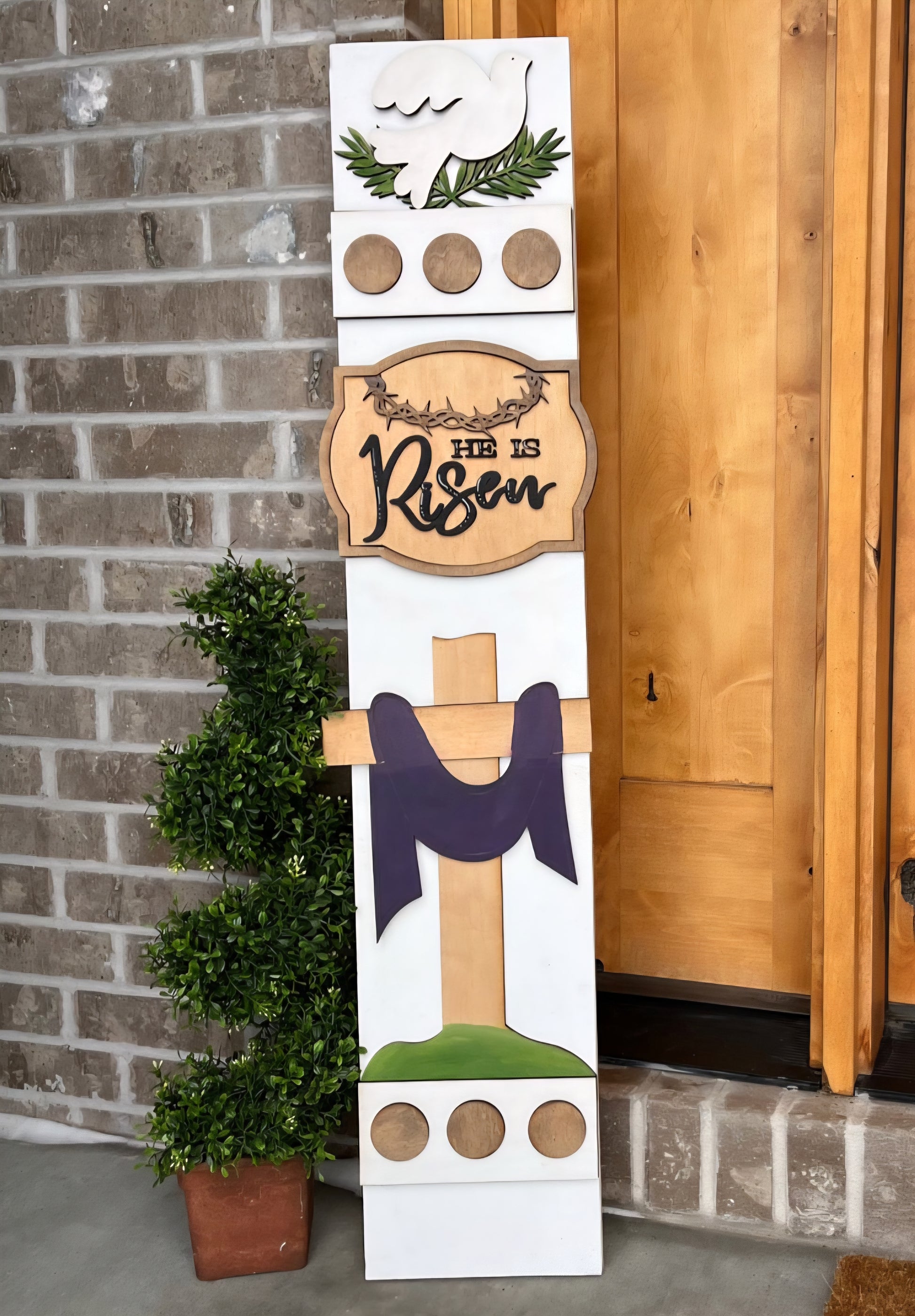Decorative Easter sign with cross, 'He is Risen' text, and greenery against a brick wall.