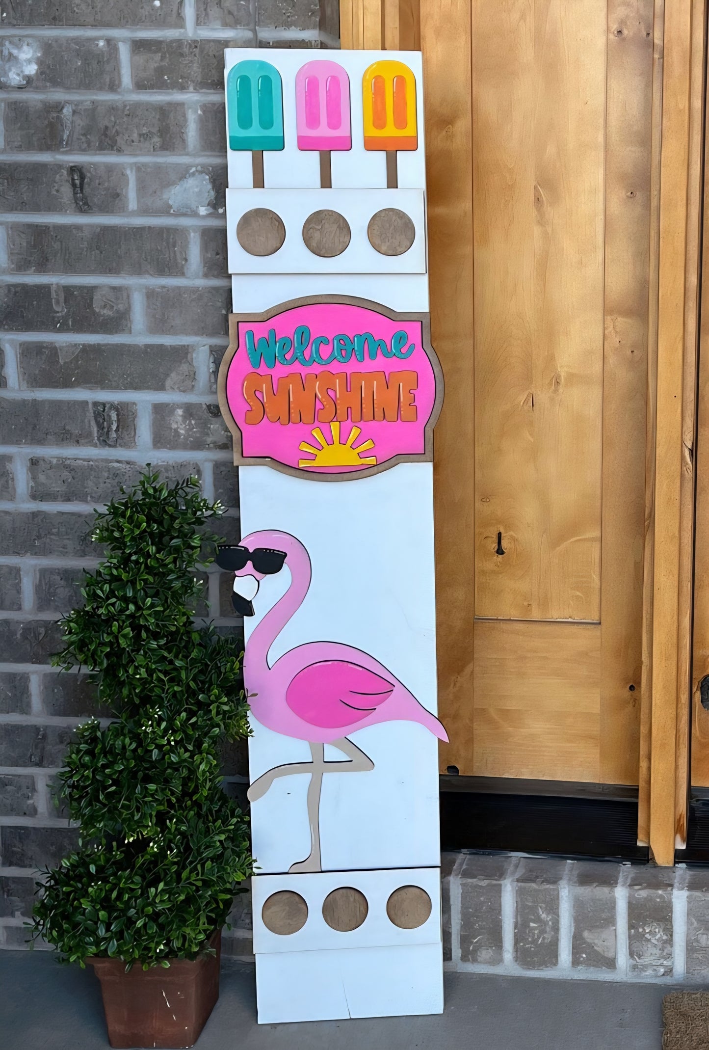 Decorative sign with colorful popsicles, 'Welcome Sunshine' text, and a pink flamingo on a brick wall background.