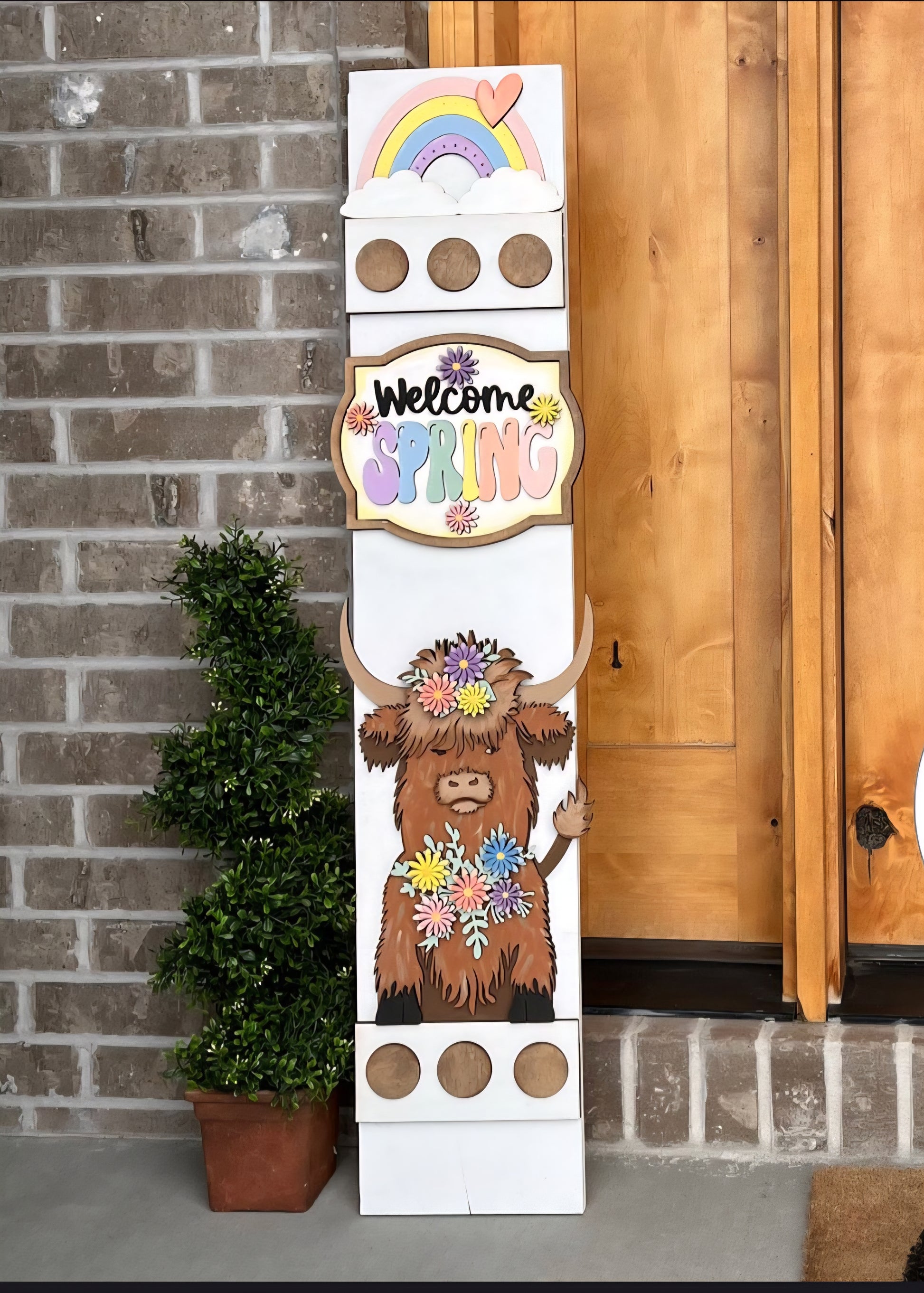 Decorative sign with a highland cow illustration and 'Welcome Spring' text against a brick wall.