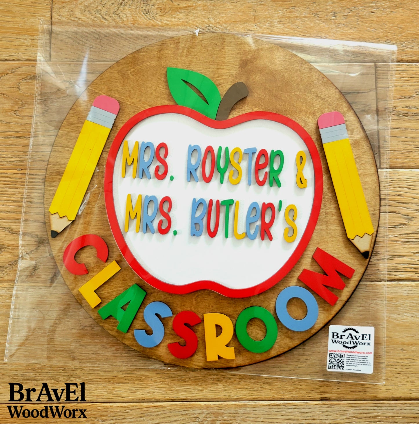 Wooden apple-shaped sign with colorful text on a wooden surface, branded with BravEl WoodWorx.