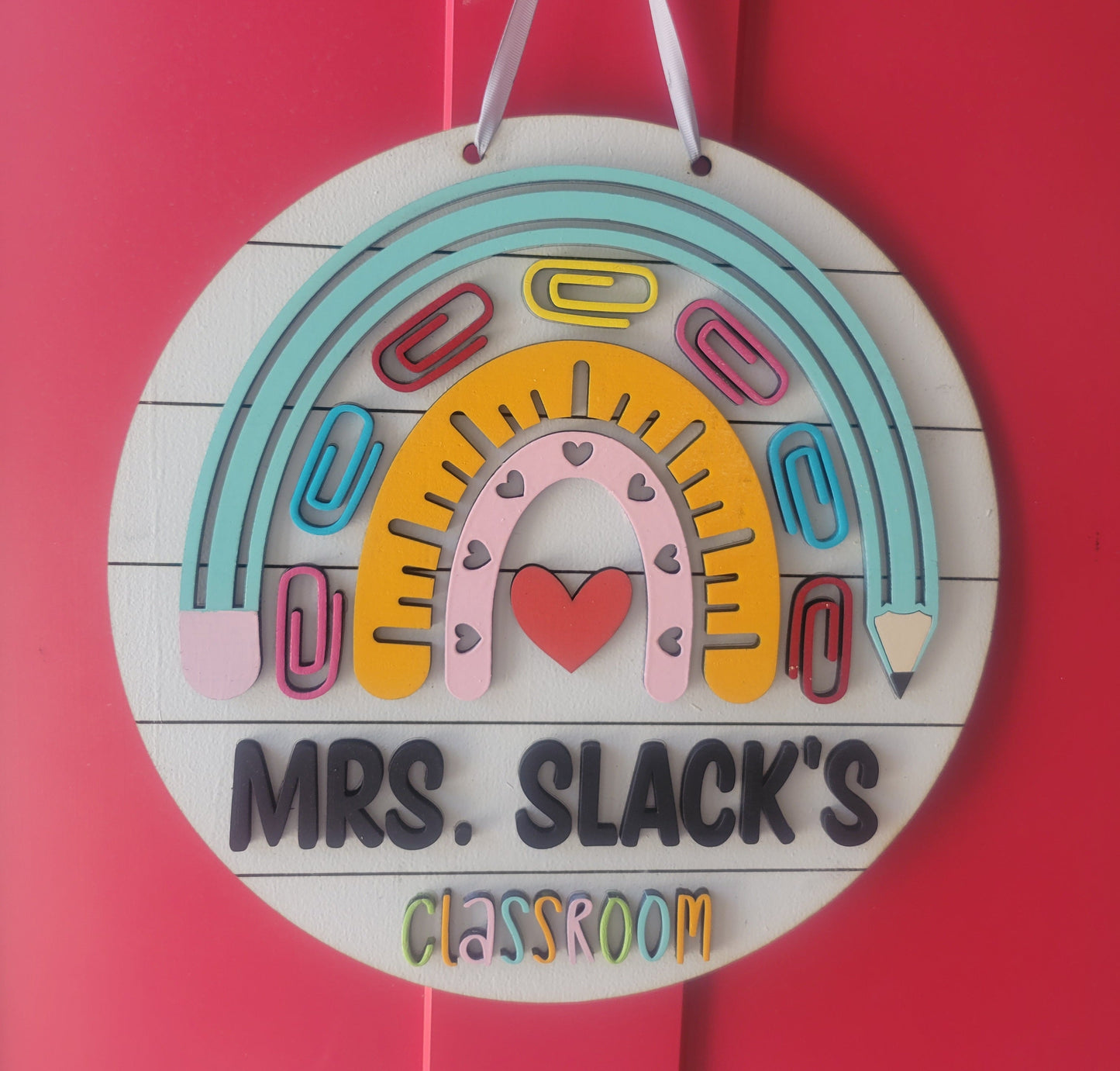 A round wooden sign with a colorful design, featuring a rainbow, hearts, and pencils, with the text 'MR. SLACK'S CLASSROOM'.