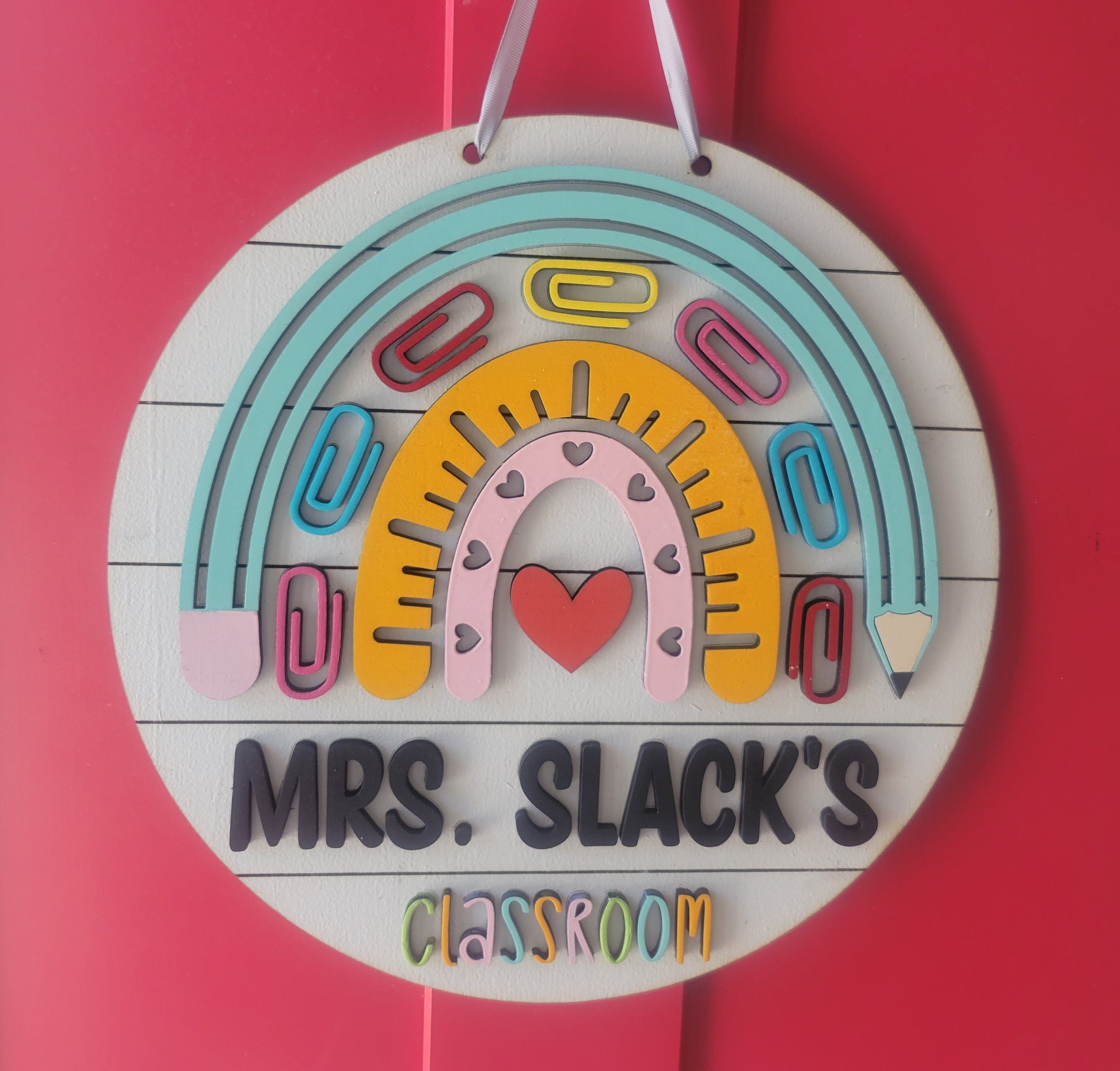 A round wooden sign with a colorful design, featuring a rainbow, hearts, and pencils, with the text 'MR. SLACK'S CLASSROOM'.